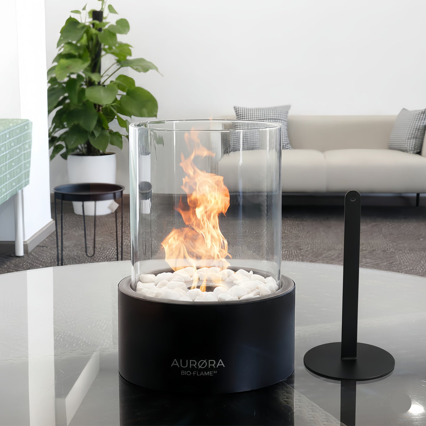 Cheminée de table au bioéthanol cylindrique de couleur noire, allumée avec une flamme vive sur un lit de galets blancs. Elle est posée sur une table en verre dans un salon moderne et lumineux, accompagnée de son accessoire éteignoir en métal noir posé à côté.