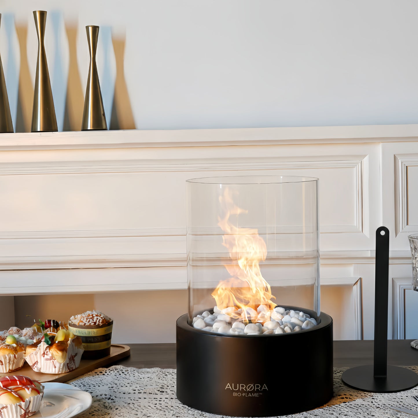 Cheminée de table au bioéthanol AURØRA BIO-FLAME™ de couleur noire, allumée avec une flamme vive sur un lit de galets blancs. Elle est présentée sur une table en bois dans une salle à manger élégante, à côté de pâtisseries et de verres à pied, avec un éteignoir noir posé à sa droite.