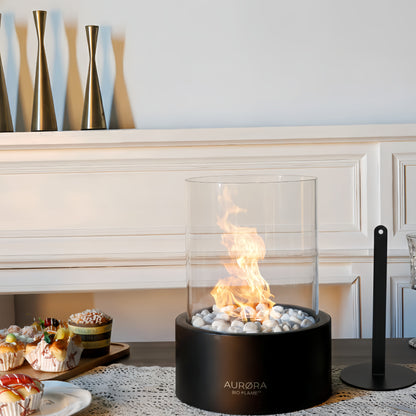 Cheminée de table au bioéthanol AURØRA BIO-FLAME™ de couleur noire, allumée avec une flamme vive sur un lit de galets blancs. Elle est présentée sur une table en bois dans une salle à manger élégante, à côté de pâtisseries et de verres à pied, avec un éteignoir noir posé à sa droite.