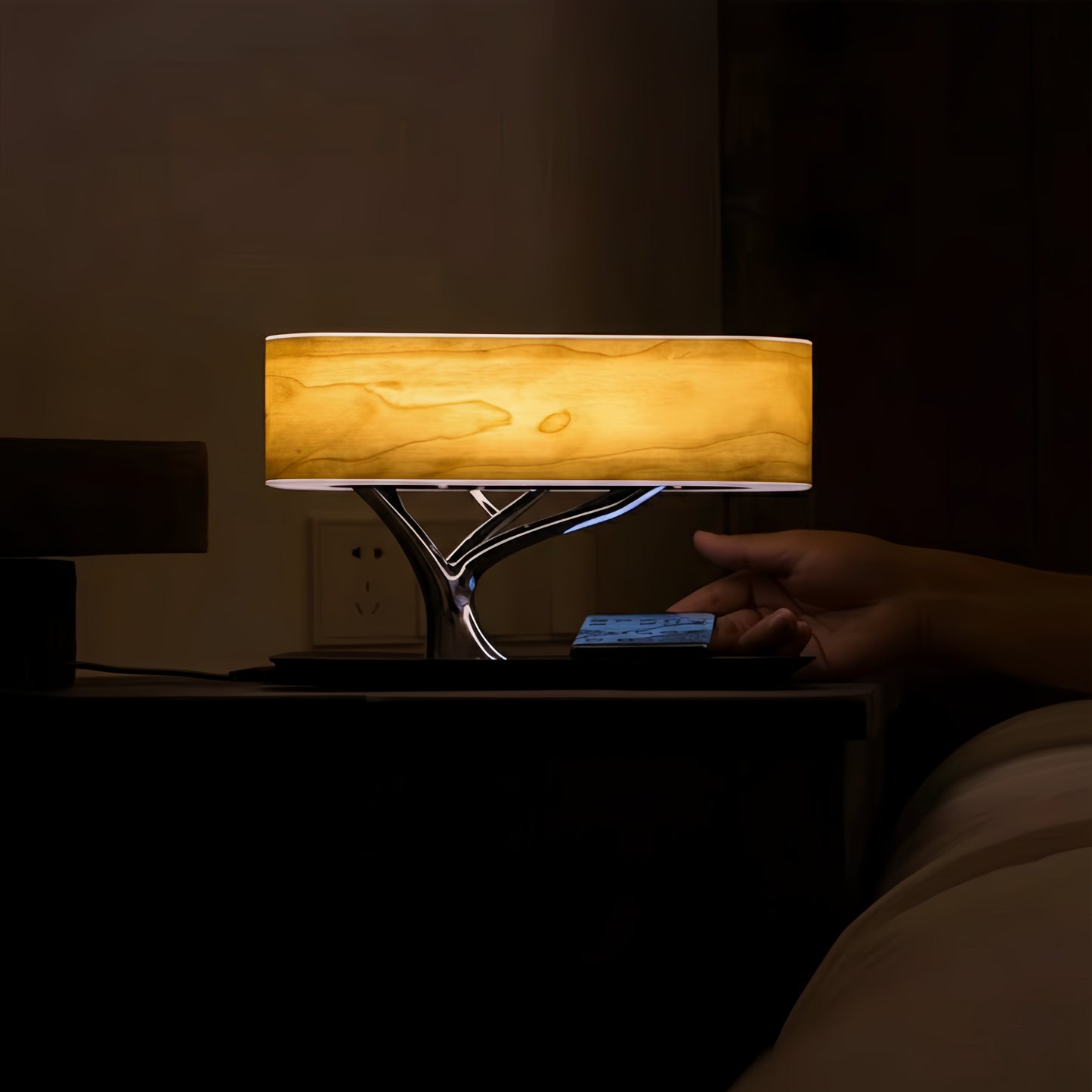 Lampe de chevet/enceinte Bluetooth design, abat-jour effet bois clair allumé, base noire, main ajustant le téléphone sur la table de nuit sombre.
